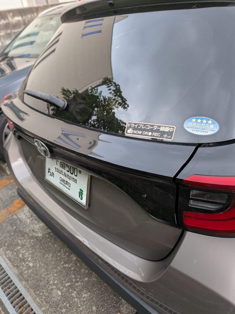 Rear view of a dark SUV with wiper on the rear window and a Japanese license plate at the bottom edge of the tailgate; two stickers on the window (a black  - 車専門鍵店カーロック 施工実績・鍵作成'NOW ON REC' label and a blue star badge).