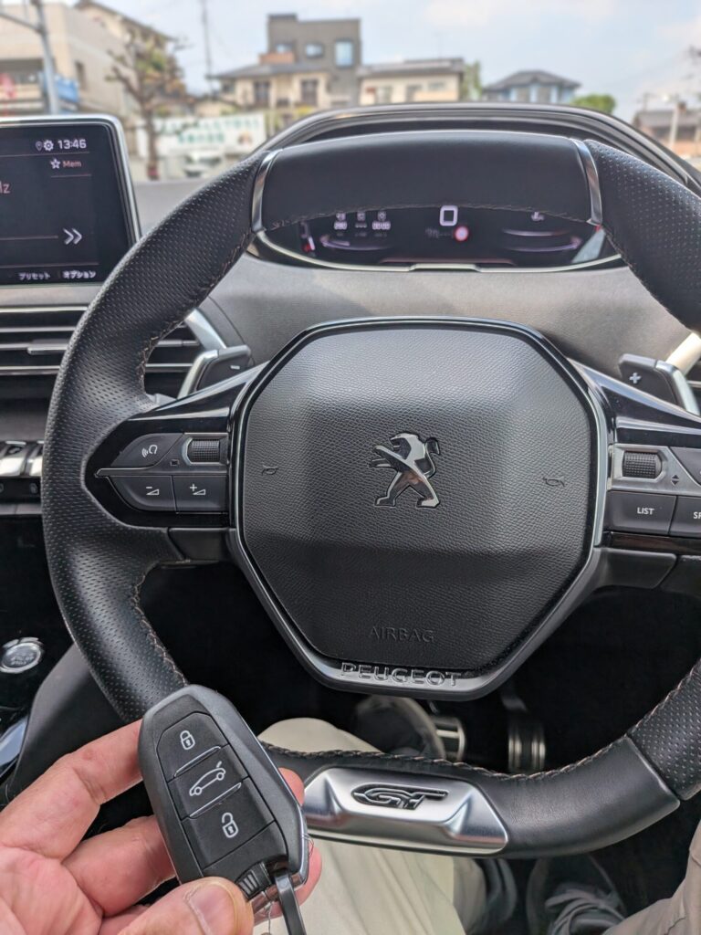 車の車内で、ダッシュボードにGTバッジが見える中、ステアリングホイールの近くでプジョーのキーフォブを手に持っている人物。 - 車専門鍵店カーロック 施工実績・鍵作成